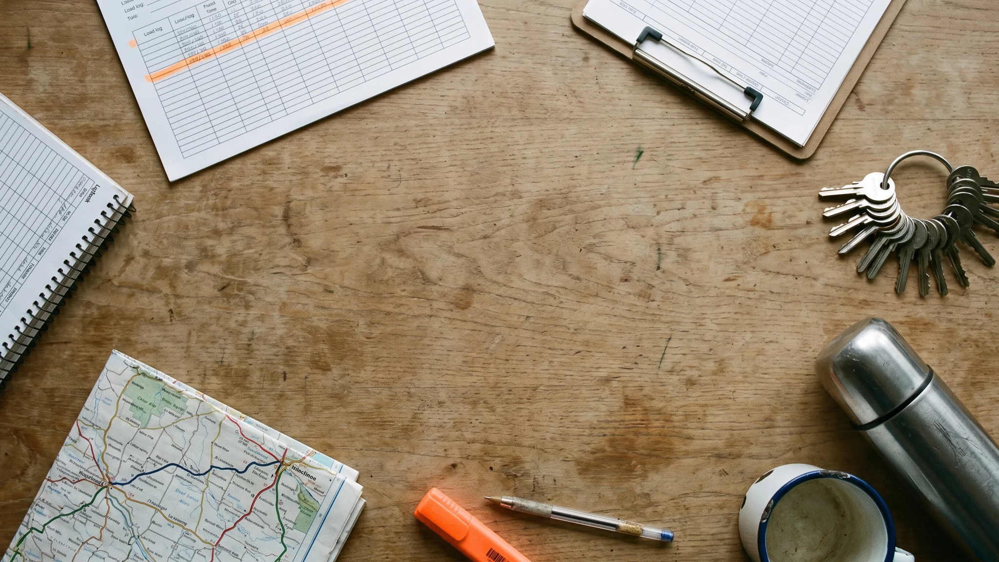 A printed load log on a wooden table, its rows struck through in orange highlighter, beside a spiral logbook, a clipboard of delivery paperwork, a bunch of trailer padlock keys, a folded road atlas and a thermos flask.